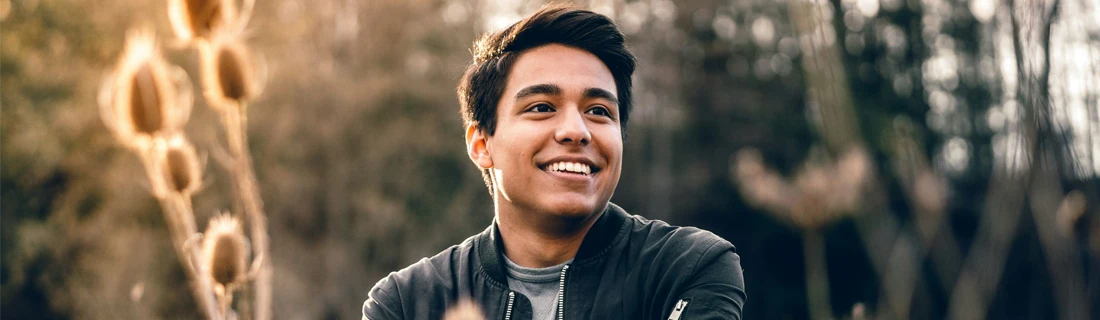 A young man smiles confidently while standing outdoors in nature, surrounded by dried plants and soft sunlight.