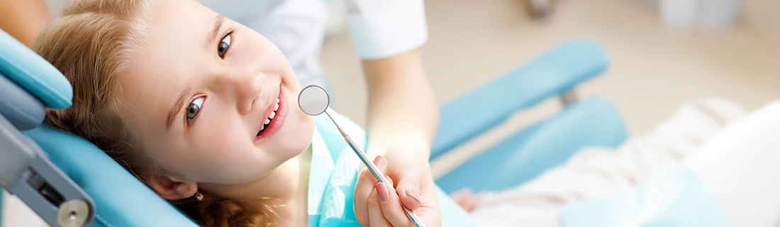 Dentist Canoga Park CA Dental Cleanings and Exams A young child with blonde hair smiles while lying in a blue dental chair as a dentist holds a mirror near the child's mouth.