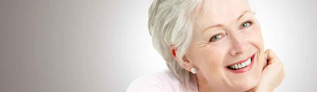 Dentist Canoga Park CA Dentures Partials A close-up shows a smiling older woman with short, gray hair and light eyes and wrinkles while resting her chin on her hand.